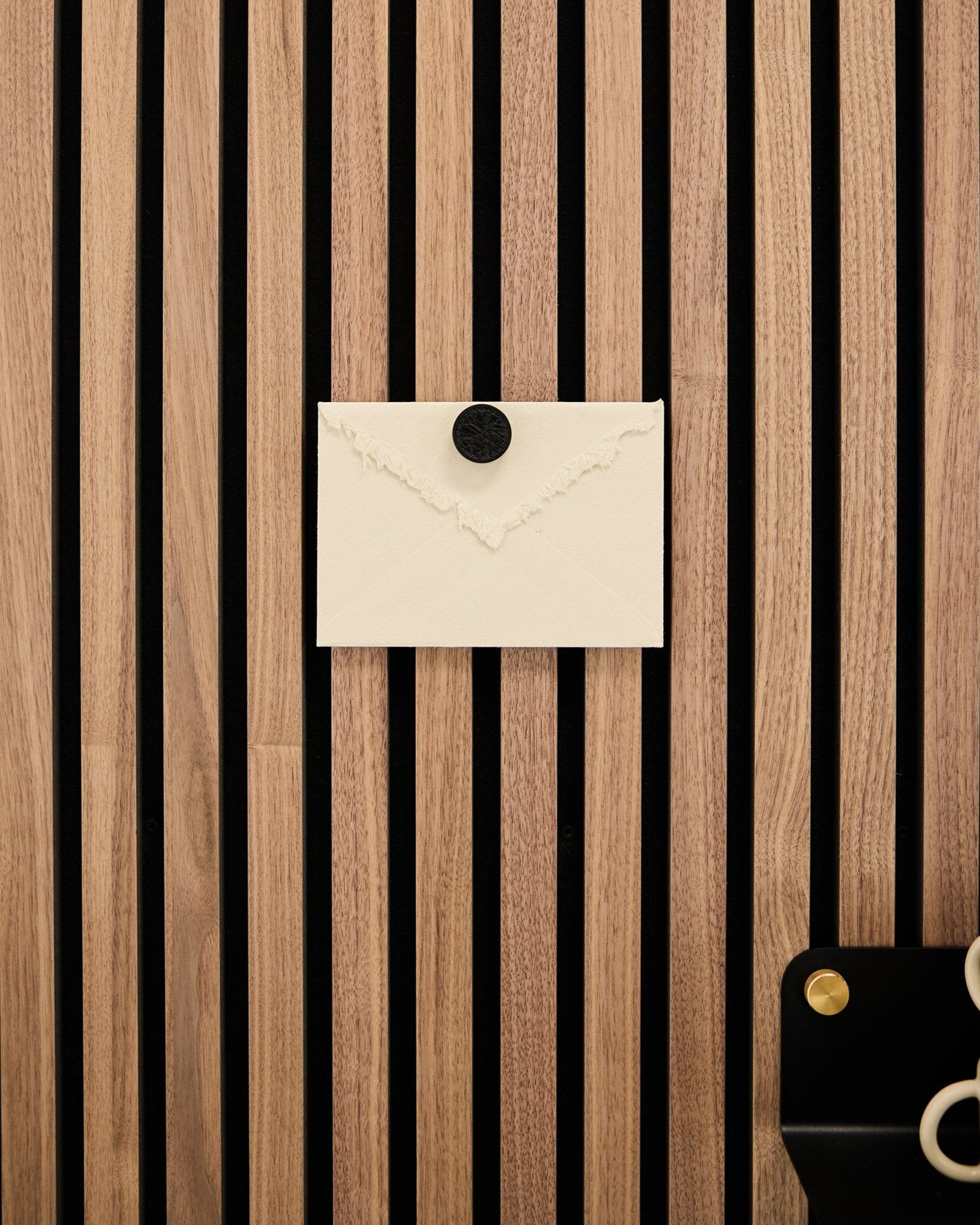 En kremfarget konvolutt er festet til et WoodUpp Akupanel eikestripet Akustikkpanel med Norr Poster Magnet-settet i Black Oak; en saks ligger nederst til høyre.
